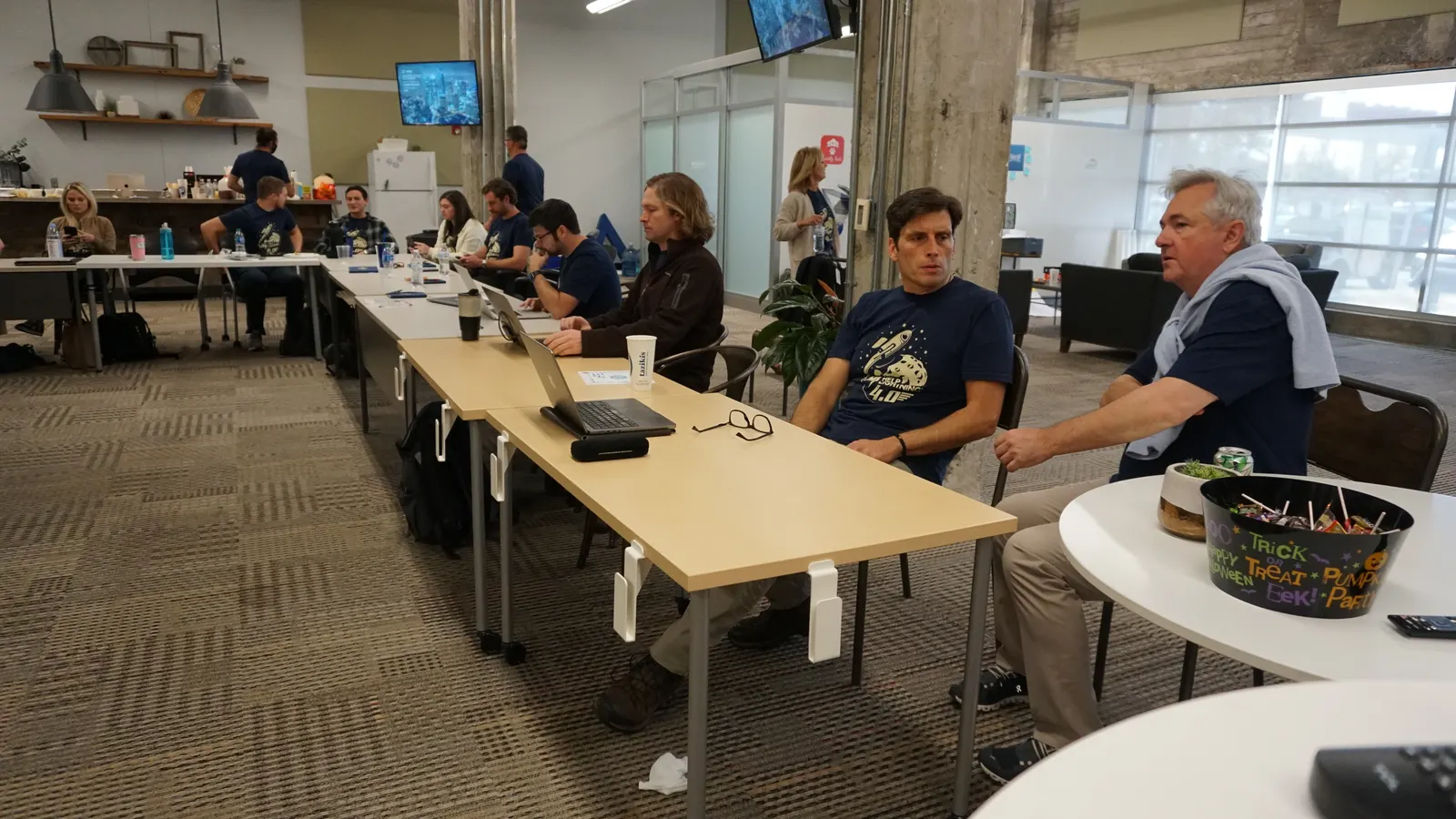 Teammates working and talking at long tables in the open office.
