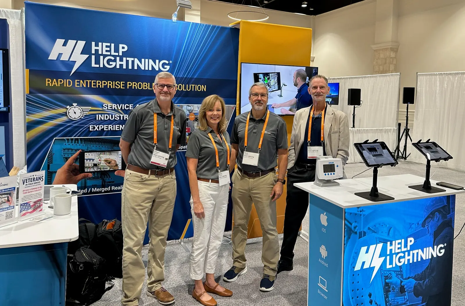 Four Help Lightning teammates at a trade-show booth with tablets and a Merged Reality demo on the counter.