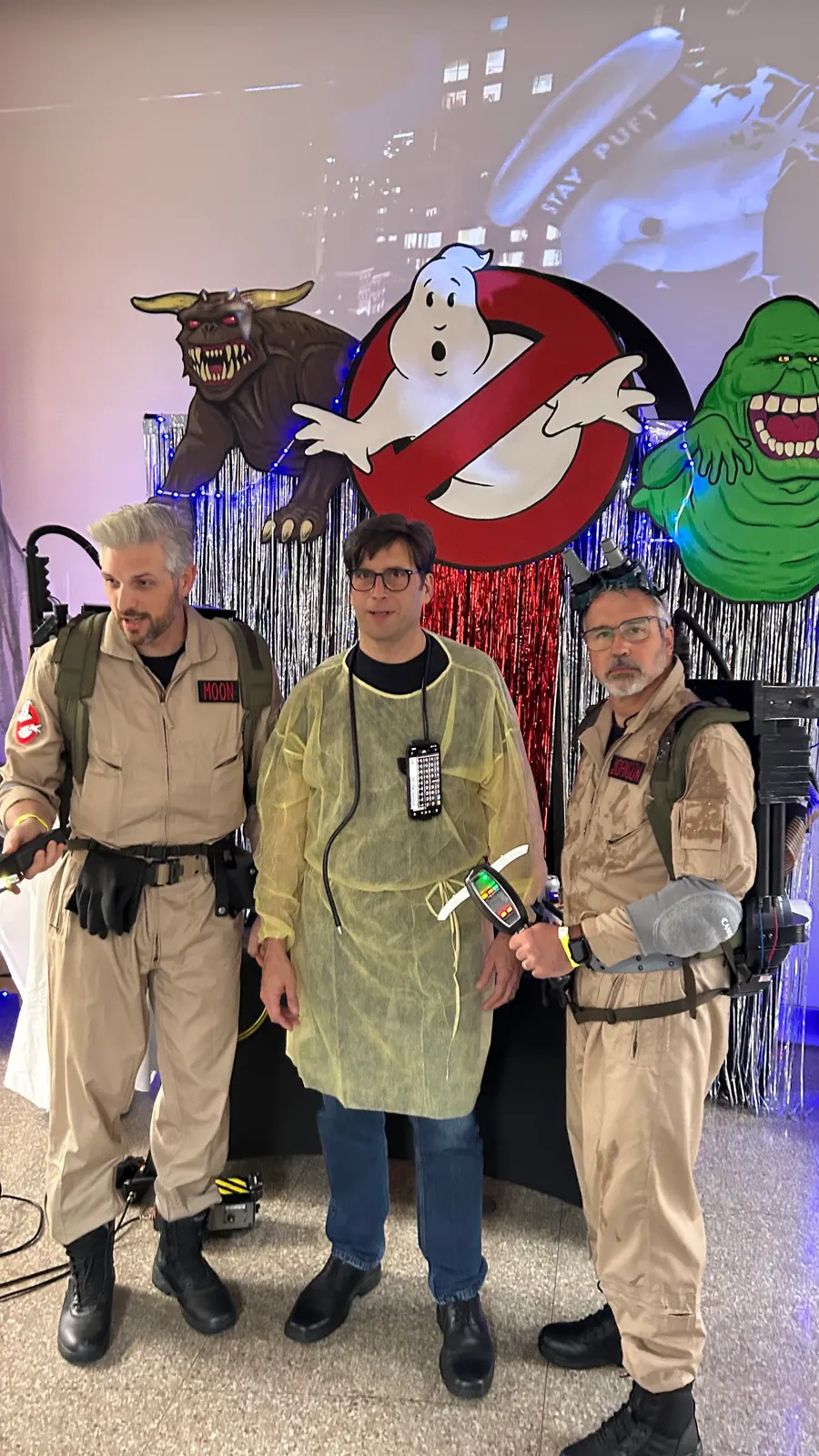 Three teammates in Halloween costumes in front of a Ghostbusters backdrop at a company party.